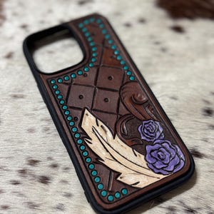 Può includere: Custodia per telefono in pelle marrone con bordo nero. La custodia presenta un design intagliato con una piuma, due rose viola e accenti color turchese. La custodia ha un motivo a diamante.