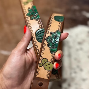 May include: Two leather belts with a light brown background and dark brown edges. The belts feature green cactus and succulent designs. The edges are decorated with a scalloped pattern. The belts are held by a person with red nail polish.