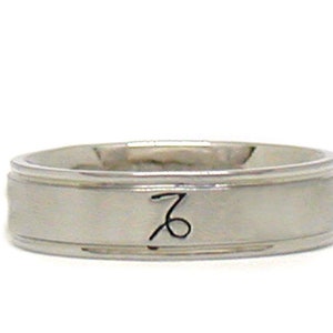 May include: A silver band ring with a simple design and a black inscription of the astrological symbol for Capricorn.