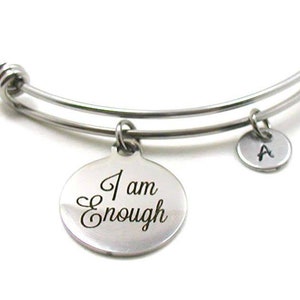 May include: Silver expandable bangle bracelet with two charms. One charm is a round disc with the text "I am Enough" and the other charm is a round disc with the letter "A".