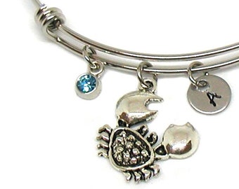 May include: Silver bangle bracelet with a crab charm, a blue crystal charm, and a silver charm with the letter 'A' engraved on it.