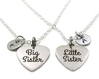 May include: Two silver necklaces with heart-shaped pendants. The pendant on the left says "Big Sister" and the pendant on the right says "Little Sister". Each necklace has a small round charm with a letter on it.