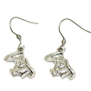 May include: A pair of silver dangle earrings featuring a small, detailed orangutan charm. The orangutan is depicted sitting with its arm raised in a salute.