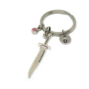 May include: Silver-toned keychain featuring a small dagger charm, a pink gemstone, and a circular charm with the letter "D". The keychain is designed with a split ring for attaching keys or other items.