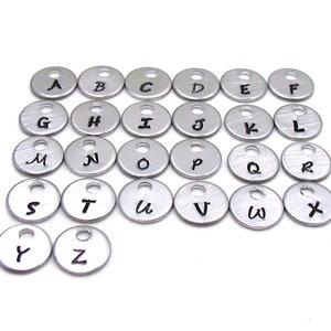 K&ouml;nnte beinhalten: Ein Satz von 26 silbernen Alphabet-Buchstabenanh&auml;ngern aus Metall. Jeder Anh&auml;nger ist ein kleiner Kreis mit einem einzelnen Buchstaben in Schwarz gedruckt.