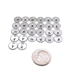 Puede incluir: Un conjunto de 26 abalorios de letras del alfabeto de metal plateado con letras negras. Los abalorios son aproximadamente del tama&ntilde;o de una moneda de 10 c&eacute;ntimos de euro.