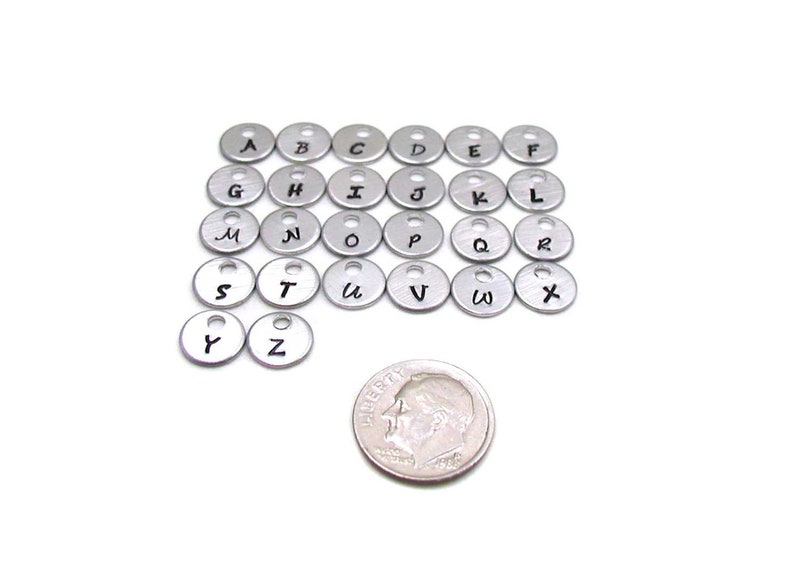Puede incluir: Un conjunto de 26 abalorios de letras del alfabeto de metal plateado con letras negras. Los abalorios son aproximadamente del tama&ntilde;o de una moneda de 10 c&eacute;ntimos de euro.