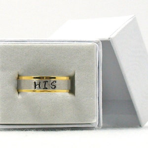 Op de afbeelding: Een zilveren ring met een gouden band en het woord "HIS" erop gegraveerd. De ring zit in een witte doos met een grijze voering.