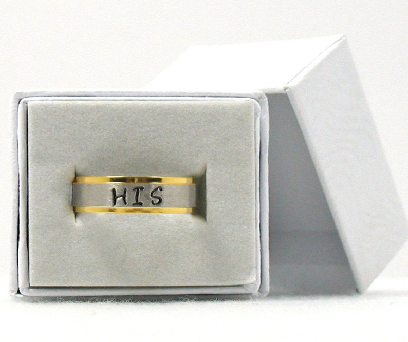 Op de afbeelding: Een zilveren ring met een gouden band en het woord "HIS" erop gegraveerd. De ring zit in een witte doos met een grijze voering.