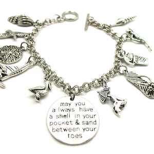 May include: Silver charm bracelet featuring a range of beach-themed charms, such as seashells, flip-flops, a sailboat, and a pelican. The bracelet also includes a round charm with the inscription "may you always have a shell in your pocket & sand between your toes".