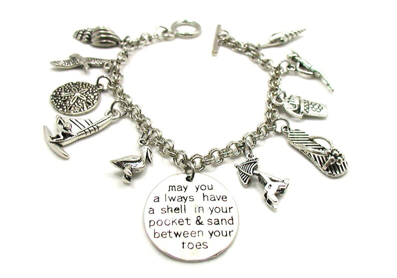 May include: Silver charm bracelet featuring a range of beach-themed charms, such as seashells, flip-flops, a sailboat, and a pelican. The bracelet also includes a round charm with the inscription "may you always have a shell in your pocket & sand between your toes".