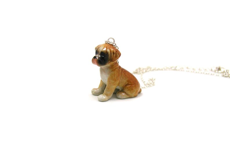 Puede incluir: Un collar con un colgante de porcelana de un perro boxer. El perro es marr&oacute;n y blanco y est&aacute; sentado con la cabeza inclinada hacia un lado. El collar es de plata.