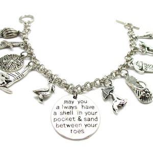 May include: Silver charm bracelet featuring a range of beach-themed charms, including a seashell, a starfish, a flip-flop, a sailboat, a pelican, and a beach umbrella. The bracelet also includes a charm with the text "may you always have a shell in your pocket & sand between your toes".