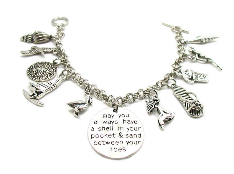 May include: Silver charm bracelet featuring a range of beach-themed charms, including a seashell, a starfish, a flip-flop, a sailboat, a pelican, and a beach umbrella. The bracelet also includes a charm with the text "may you always have a shell in your pocket & sand between your toes".