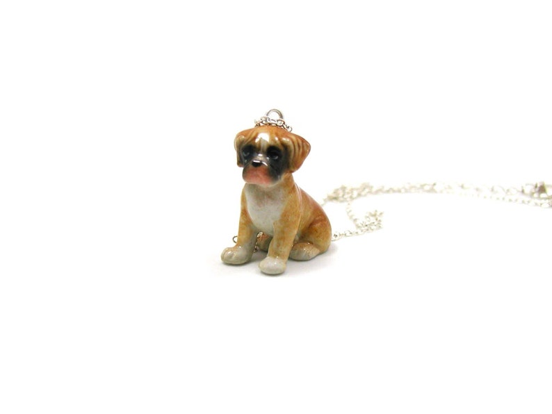 Puede incluir: Un collar con un colgante de porcelana que representa a un perro boxer marr&oacute;n y blanco. El perro est&aacute; sentado con la cabeza ligeramente inclinada hacia un lado. El collar est&aacute; hecho de cadena de plata.