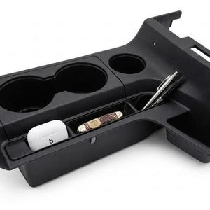 May include: A black plastic automotive center console organizer featuring two circular cup holders and a long, narrow storage tray. The tray contains a white wireless earbud case, a folding pocket knife with a wood handle, and two pens.