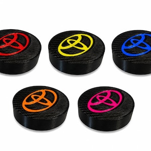 May include: Five circular black emblems with a carbon fiber texture, each featuring a raised Toyota logo in a different color: red, yellow, blue, orange, and pink. These round automotive accessories have a flat top and a smooth, thick side profile.
