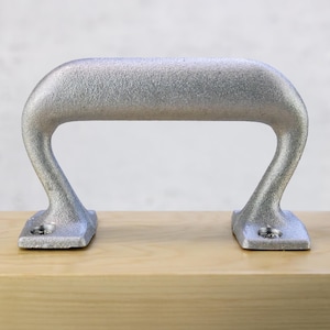 May include: A silver-colored, arched handle with a textured surface. The handle has a rectangular base on each end, designed for mounting. The handle is made of metal and is likely for a cabinet or drawer.