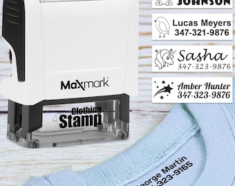 May include: A white Maxmark clothing stamp with black accents, shown with examples of stamped labels. The labels include names like Michael Johnson, Sasha, and Amber Hunter, along with phone numbers. The stamp is shown on a light blue garment.