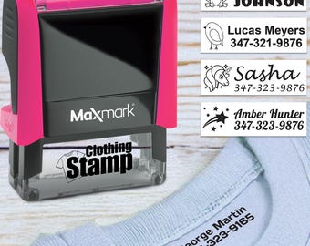 May include: A pink and black Maxmark clothing stamp with the words "Clothing Stamp" printed on the bottom. The image also shows examples of stamped labels with names and phone numbers, including "Michael Johnson" and "Sasha".