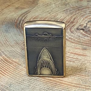 May include: A gold-colored metal lighter with a shark design. The front of the lighter features a detailed engraving of a shark's open mouth, with a swimmer above. The lighter has a rectangular shape.