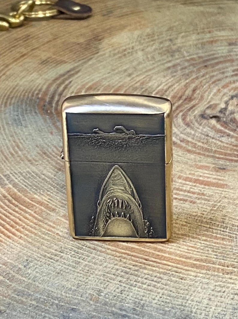 May include: A gold-colored metal lighter with a shark design. The front of the lighter features a detailed engraving of a shark's open mouth, with a swimmer above. The lighter has a rectangular shape.