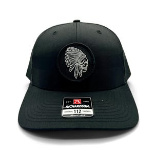 May include: Black trucker hat with a white embroidered skull wearing a headdress. The hat has a black mesh back and a white label that reads "EST. 1970 RICHARDSON. ORIGINAL 112 TRUCKER ADJUSTABLE".