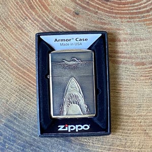 May include: A Zippo Armor Case lighter with a shark design. The lighter is in a black box and features a detailed engraving of a shark's open mouth. The text "Armor Case" and "Made in USA" are visible on the box.
