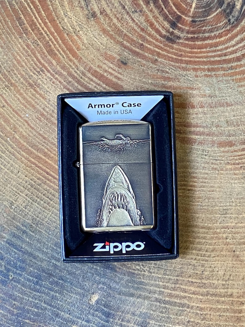 May include: A Zippo Armor Case lighter with a shark design. The lighter is in a black box and features a detailed engraving of a shark's open mouth. The text "Armor Case" and "Made in USA" are visible on the box.