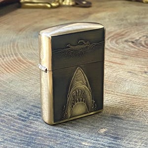 May include: A brass-colored lighter with a shark design. The front of the lighter features a detailed engraving of a shark's open mouth, with a swimmer above. The lighter has a hinged lid and a metallic finish.