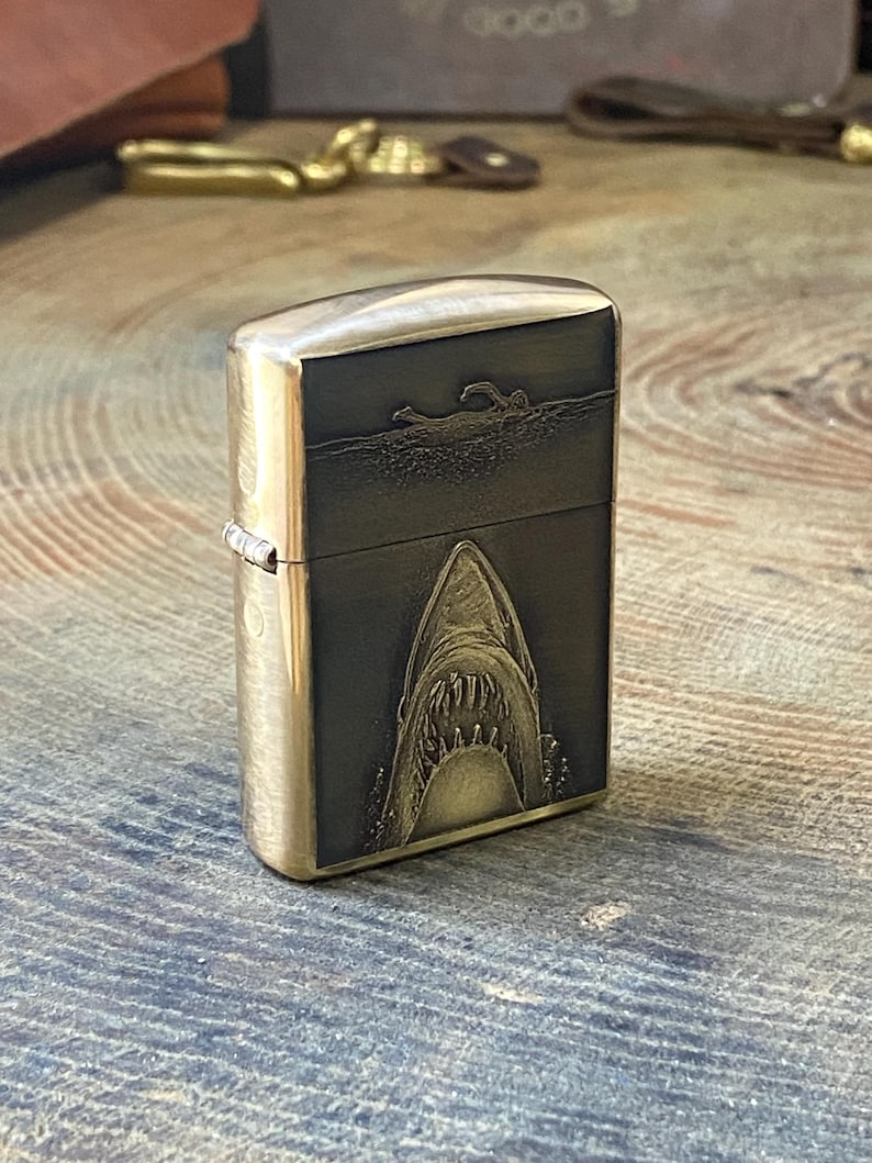 May include: A brass-colored lighter with a shark design. The front of the lighter features a detailed engraving of a shark's open mouth, with a swimmer above. The lighter has a hinged lid and a metallic finish.