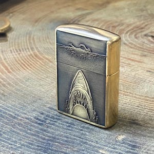 May include: A brass-colored lighter with a detailed shark design. The front of the lighter features a swimmer in the water above a shark's open mouth. The lighter has a rectangular shape with rounded edges.