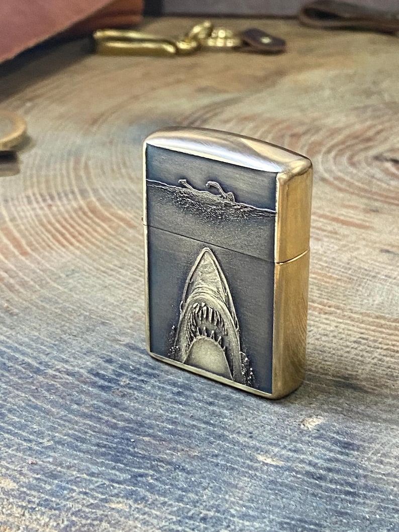 May include: A brass-colored lighter with a detailed shark design. The front of the lighter features a swimmer in the water above a shark's open mouth. The lighter has a rectangular shape with rounded edges.