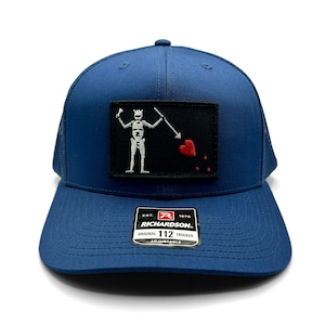May include: A navy blue trucker hat with a black patch showing a white skeleton holding a spear and a red heart. The hat has a curved peak and a mesh back. The Richardson brand label is visible.