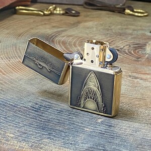 May include: A brass-colored lighter with an open lid. The front of the lighter features a detailed engraving of a shark's open mouth. The lid has a textured design. The lighter is on a wooden surface.