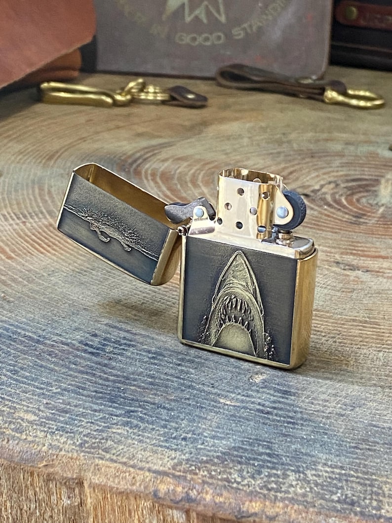 May include: A brass-colored lighter with an open lid. The front of the lighter features a detailed engraving of a shark's open mouth. The lid has a textured design. The lighter is on a wooden surface.