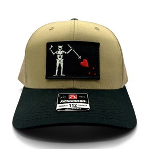 May include: A tan and black Richardson 112 trucker hat. The front panel is black, with a patch featuring a white skeleton holding a spear and a red heart. The hat has a curved brim and an adjustable strap.
