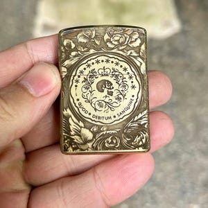 May include: A brass-colored lighter with an engraved floral and circular design. The circular design features a skull and crown motif with the Latin phrase "Quod Debitum Sanguine". The lighter is held in a hand.