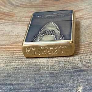 May include: A rectangular, brass-colored lighter with a detailed shark design. The top of the lighter features a shark emerging from the water, with the text "Zippo" and "BRADFORD, PA. MADE IN USA" engraved on the bottom.