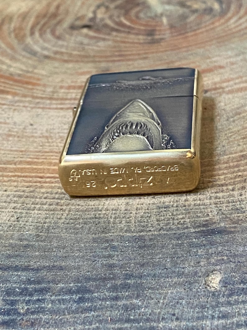 May include: A rectangular, brass-colored lighter with a detailed shark design. The top of the lighter features a shark emerging from the water, with the text "Zippo" and "BRADFORD, PA. MADE IN USA" engraved on the bottom.