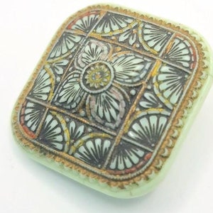 May include: A square, green button with a gold, black, and white floral design.
