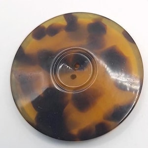 Könnte beinhalten: Ein großer, runder, brauner und schwarzer Knopf mit einem Schildpattmuster. Der Knopf hat zwei Löcher in der Mitte.