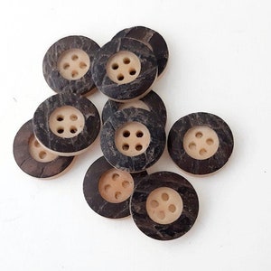Può includere: Un gruppo di dieci bottoni rotondi di colore marrone e crema, ognuno con quattro fori. I bottoni sono realizzati con materiali naturali e hanno un aspetto rustico.