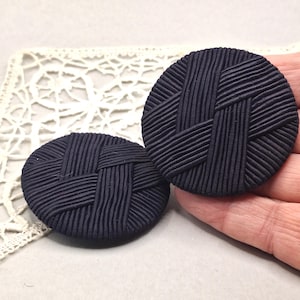 Può includere: Due bottoni rotondi blu navy con una trama intrecciata. I bottoni sono realizzati in un materiale scuro e solido e presentano un motivo a coste che crea un design a intreccio. I bottoni sono su uno sfondo neutro.