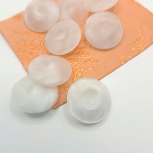 Może przedstawiać: Grupa białych, okrągłych, plastikowych guzików z fakturowaną powierzchnią. Guziki są ułożone na brzoskwiniowym materiale.