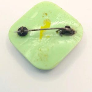 May include: A green square-shaped pin with a yellow design and a silver pin back.