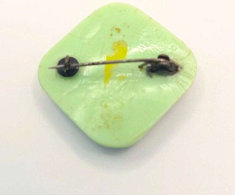 May include: A green square-shaped pin with a yellow design and a silver pin back.
