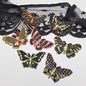 May include: A collection of six embroidered butterfly patches in various color combinations including orange, pink, green, and purple, all featuring black outlines and intricate wing details, arranged below a strip of black floral lace trim.