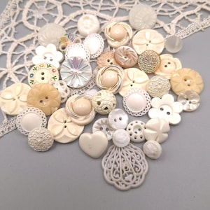 Puede incluir: Una colección de botones variados en diversos tonos de blanco, crema y beige. Los botones presentan texturas diversas, incluyendo diseños lisos, facetados, florales, en forma de corazón y de filigrana, algunos con detalles metálicos.