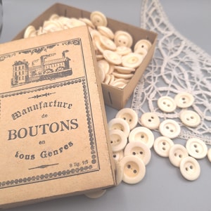 Può includere: Una scatola di cartone riempita con piccoli bottoni rotondi color bianco sporco, con un'etichetta in stile vintage che riporta la scritta "Manufacture de BOUTONS en tous Genres" e "8 lig. 1/2". Diversi bottoni sono sparsi fuori dalla scatola.